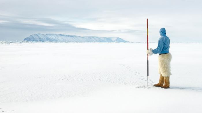 Photograph showing an indigenous man holding a spear and standing on a sea of ice looking out at snow capped mountains.