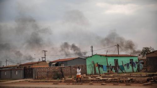 A man walks in front of a buildings, with smoke rising in the background