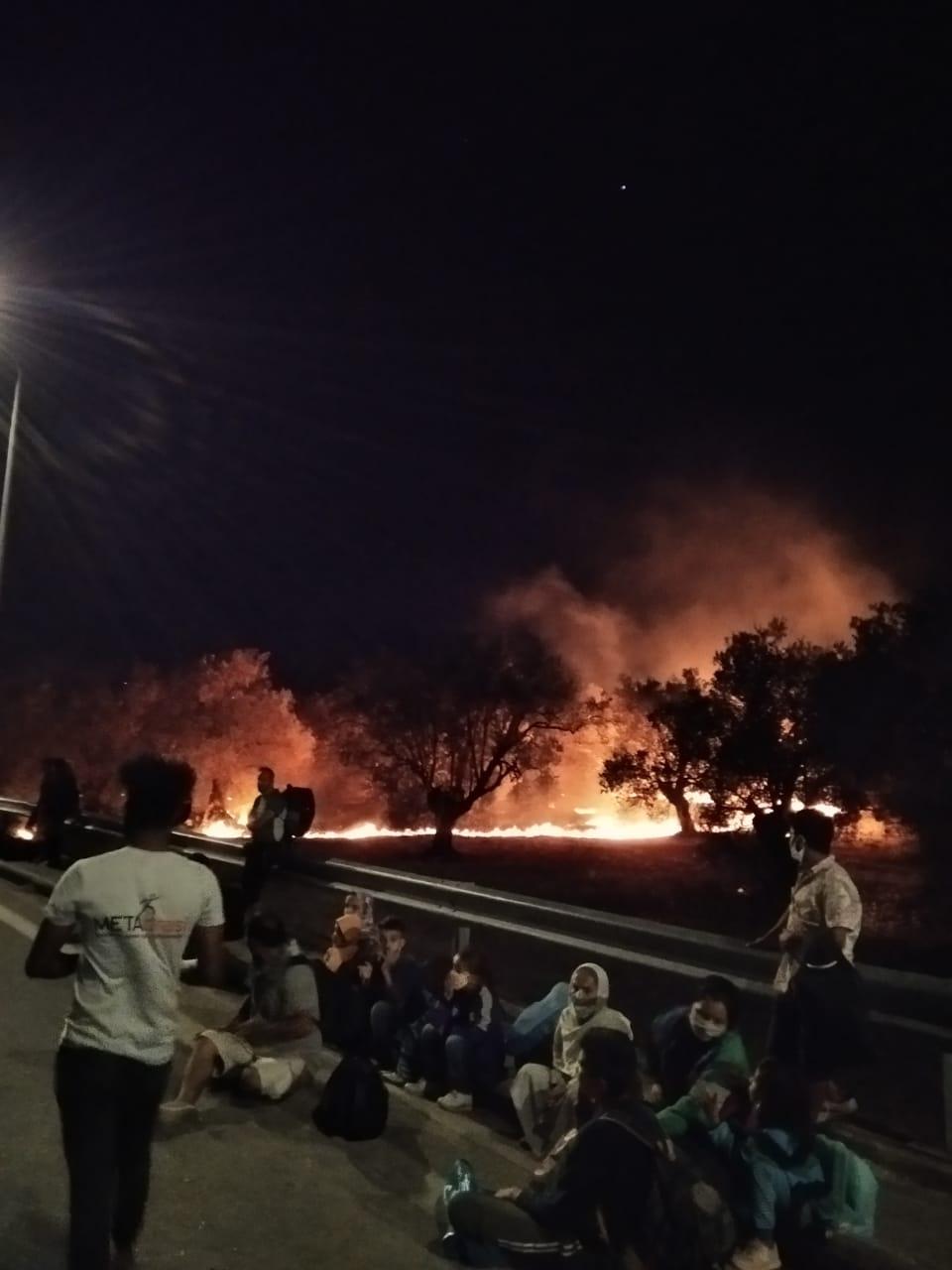 People sit on a roadside while a fire burns behind them