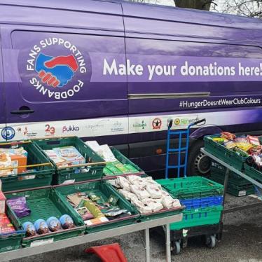 A mobile food pantry with food collected by Fans Supporting Foodbanks and other local anti-poverty groups set up at at St. Mary’s Millennium Centre, Liverpool, UK. Photo shows a purple van with cases of food set up in front of it.